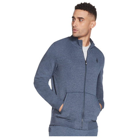 A man is standing with his hands in the pockets of a blue zip-up hoodie he appears thoughtful while looking to the side in a neutral background