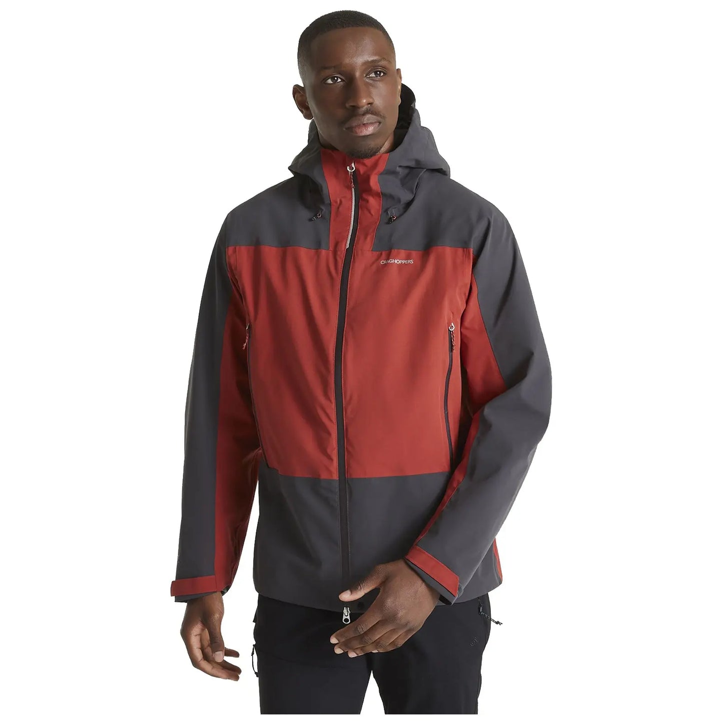 A man is standing while wearing a red and gray outdoor jacket with a hood he holds his left hand near the jacket's zipper against a plain white background.