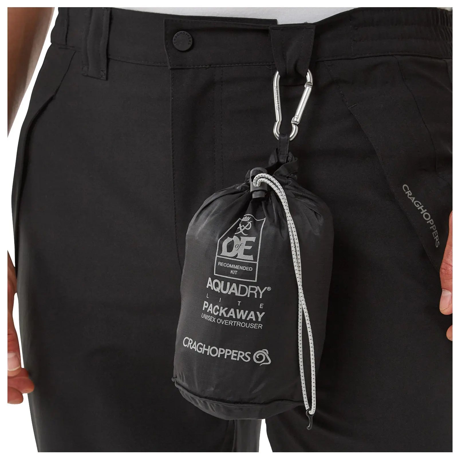 A black lightweight overtrouser is being held by a person wearing dark pants the overtrouser is packaged in a compact bag with a drawstring and a carabiner for attachment