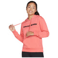 A woman wearing a bright pink sweatshirt with the word SKECHERS prominently displayed on the front smiles while holding the hoodie drawstring in one hand in a plain background