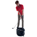 A man stands on a black training bag preparing to swing a golf club with a focused expression wearing a red shirt and gray pants in a brightly lit environment.