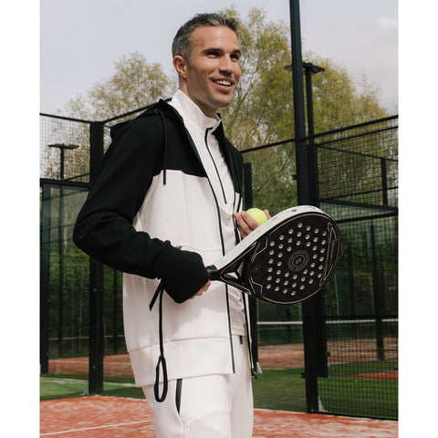 Robin van Persie wearing a black and white athletic outfit holds a padel racket and a tennis ball, smiling on a padel court surrounded by green trees and a mesh fence.