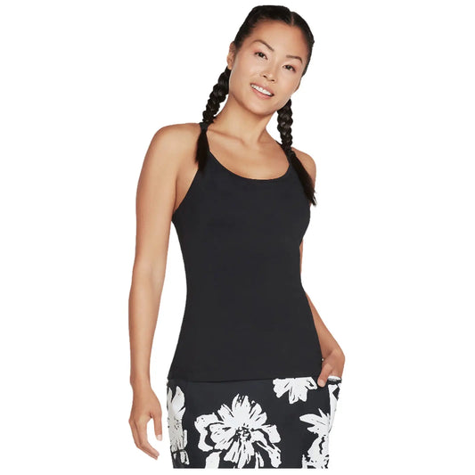 A woman wearing a black tank top and floral-patterned skirt stands smiling with her hands on her hips in a bright environment suggesting a casual or athletic setting.