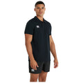 A man in a black polo shirt and shorts stands facing the viewer with a neutral expression in a plain background showcasing athletic apparel.