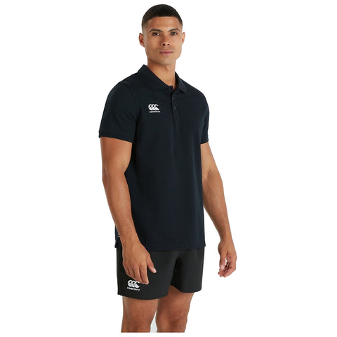 A man in a black polo shirt and shorts stands facing the viewer with a neutral expression in a plain background showcasing athletic apparel.