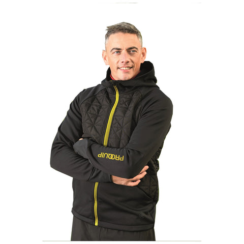 A man is smiling while wearing a zip-up, black hoodie with a quilted front and a yellow logo on the sleeve in a plain white background.