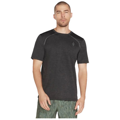 A man stands wearing a short-sleeve black athletic shirt and patterned green shorts with a neutral expression against a plain white background. He appears ready for exercise or casual wear.