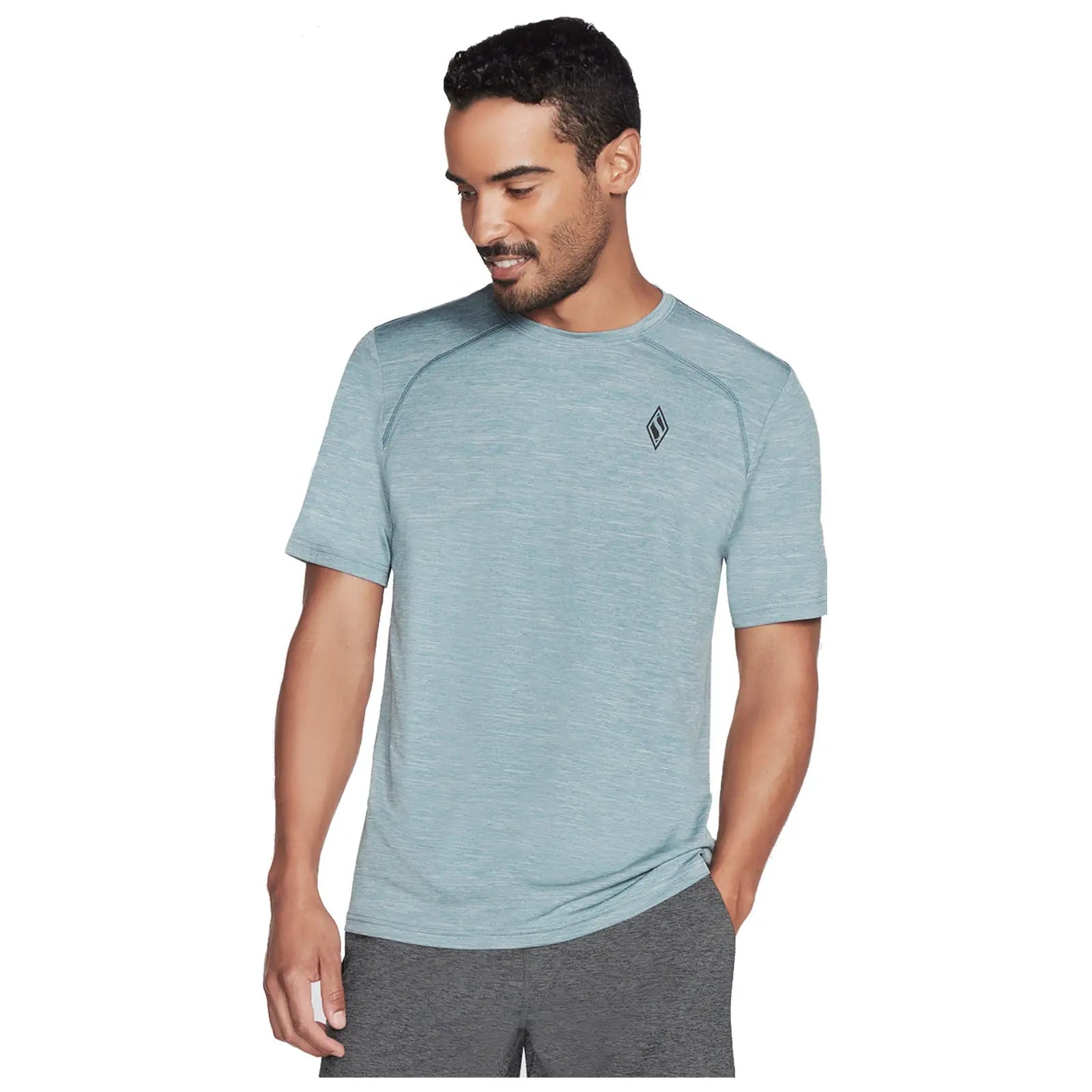 A man in a light blue athletic shirt stands smiling while wearing grey shorts in a neutral-background setting, conveying a casual and active appearance.