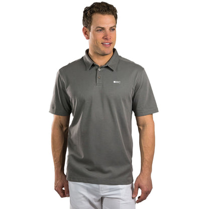 A man stands smiling wearing a dark gray polo shirt and white pants in a neutral background, conveying a casual and relaxed appearance.
