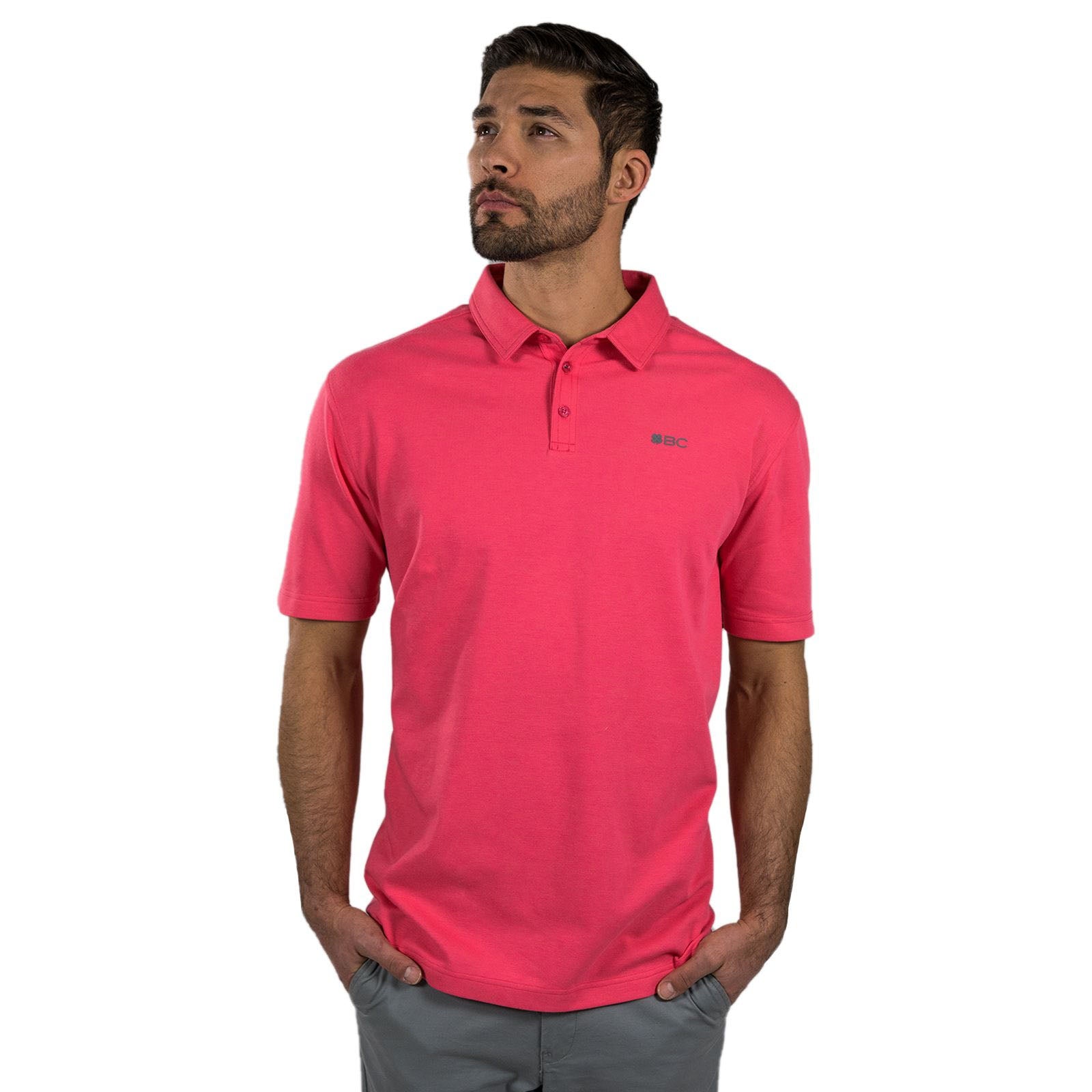 A man stands confidently wearing a pink polo shirt with short sleeves and a collar while looking upward with his hands placed casually in his pockets against a plain white background.