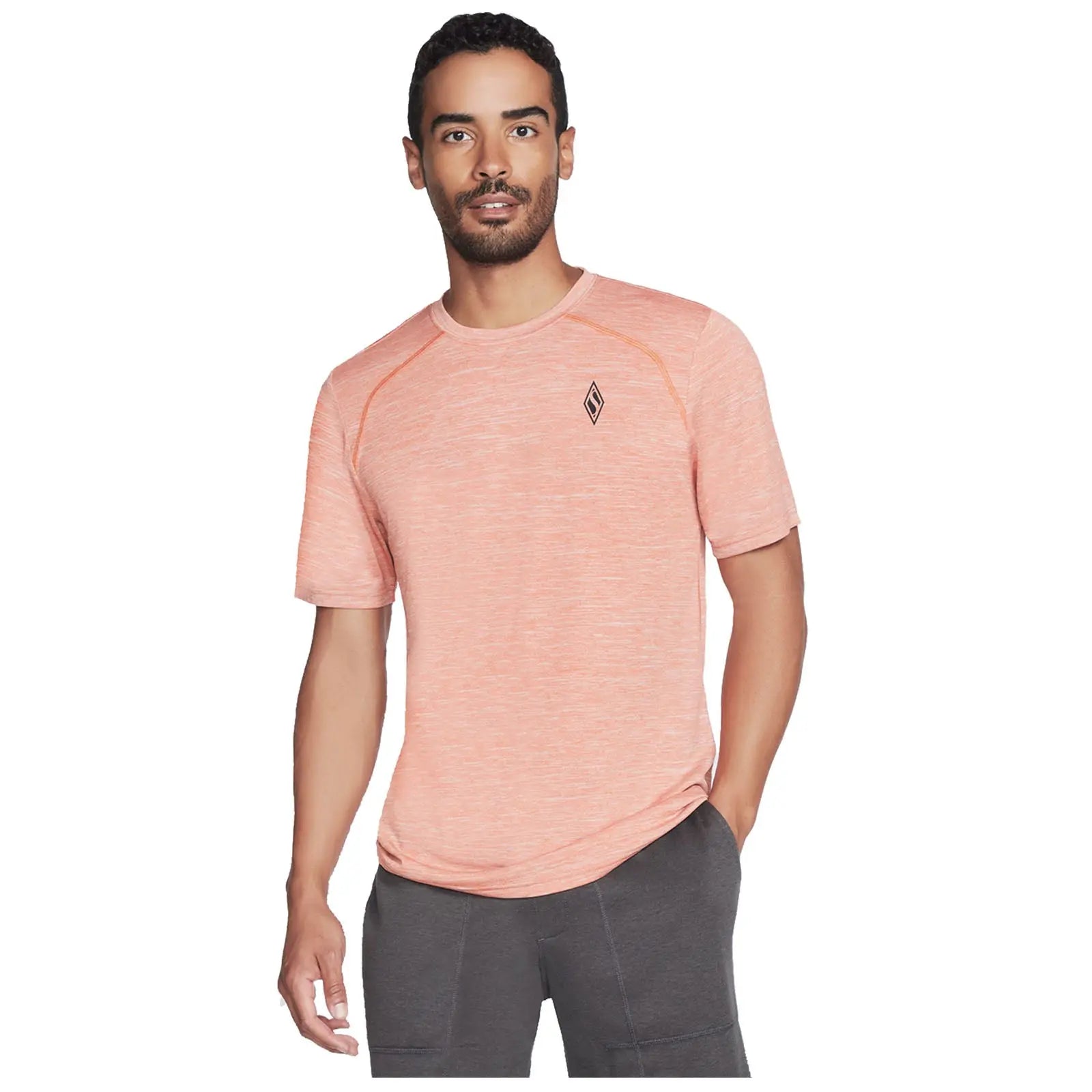 A man is standing and smiling while wearing a light orange athletic t-shirt and dark gray shorts in a neutral background, showcasing casual sporty attire.