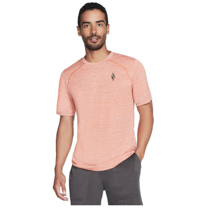 A man is standing and smiling while wearing a light orange athletic t-shirt and dark gray shorts in a neutral background, showcasing casual sporty attire.