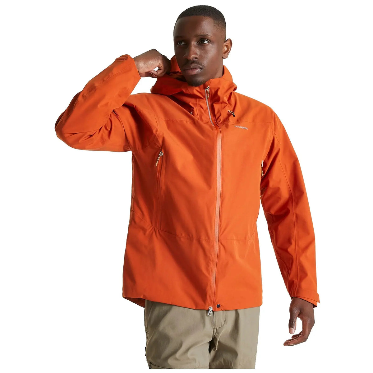 A person wears an orange jacket and poses with one hand raised near their face while looking directly ahead in a neutral background.