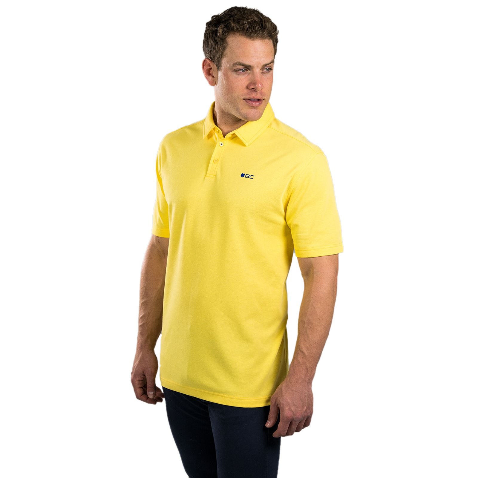 A man stands wearing a yellow polo shirt with a small logo on the left side, facing slightly to the right in a plain white background, looking confidently off to the side.
