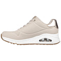 A beige athletic shoe with a shiny gold accent on the heel features a cushioned white sole and an air pocket visible on the side designed for comfort and support.