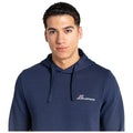 A young man is standing and looking at the camera while wearing a navy blue hoodie with a logo on the chest in a neutral background.