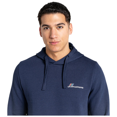 A young man is standing and looking at the camera while wearing a navy blue hoodie with a logo on the chest in a neutral background.