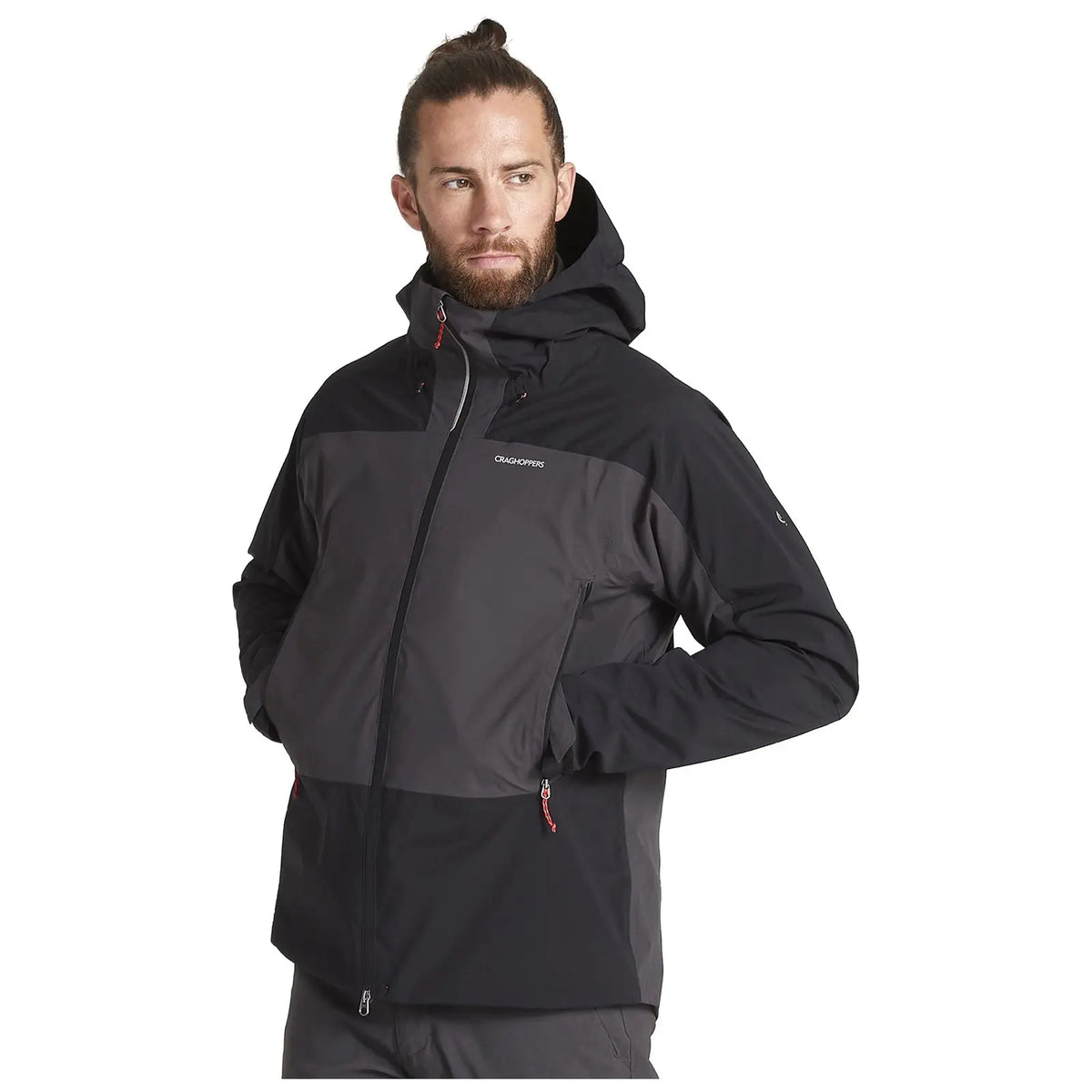 A man stands confidently wearing a black and gray ski jacket with a hood. He has long hair tied back and a thoughtful expression in a neutral environment.