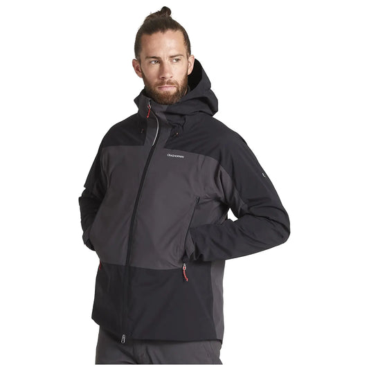 A man stands confidently wearing a black and gray ski jacket with a hood. He has long hair tied back and a thoughtful expression in a neutral environment.