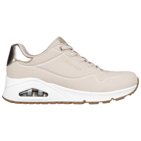 A beige sneaker with a metallic gold accent is displayed side-on featuring perforations on the upper part and a visible air cushioning unit in the white midsole