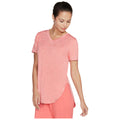 A woman wears a pink short-sleeve V-neck shirt and pink pants while looking down at her shirt with a slight smile in a neutral background.