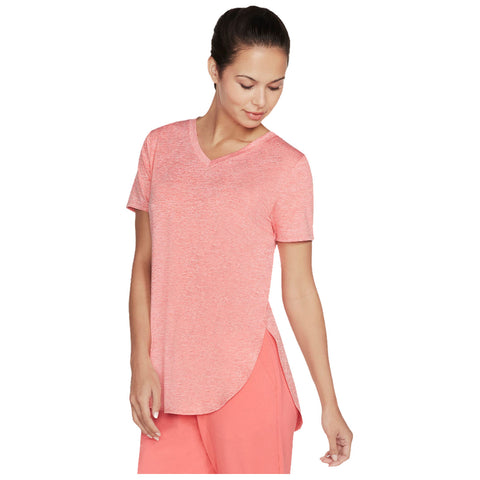A woman wears a pink short-sleeve V-neck shirt and pink pants while looking down at her shirt with a slight smile in a neutral background.