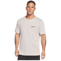 A man wearing a light-colored t-shirt with a small logo stands straight facing the viewer in a plain white background, presenting a casual and relaxed appearance.