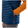 A person wearing a blue puffer vest places their hand inside a pocket while wearing an orange long-sleeve shirt in a neutral background.