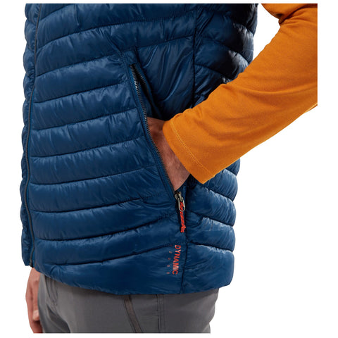 A person wearing a blue puffer vest places their hand inside a pocket while wearing an orange long-sleeve shirt in a neutral background.