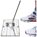 A golf club is poised above a golf ball resting on a putting mat with alignment guides while a white athletic shoe is positioned next to the mat on the right side.