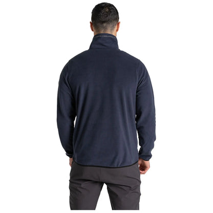 A man is standing with his back facing the viewer wearing a dark blue fleece jacket and dark pants in a neutral background suggesting an indoor setting.