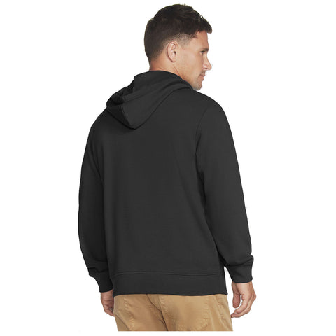 A man is standing with his back facing the viewer wearing a black hoodie and light-colored pants in a neutral background.