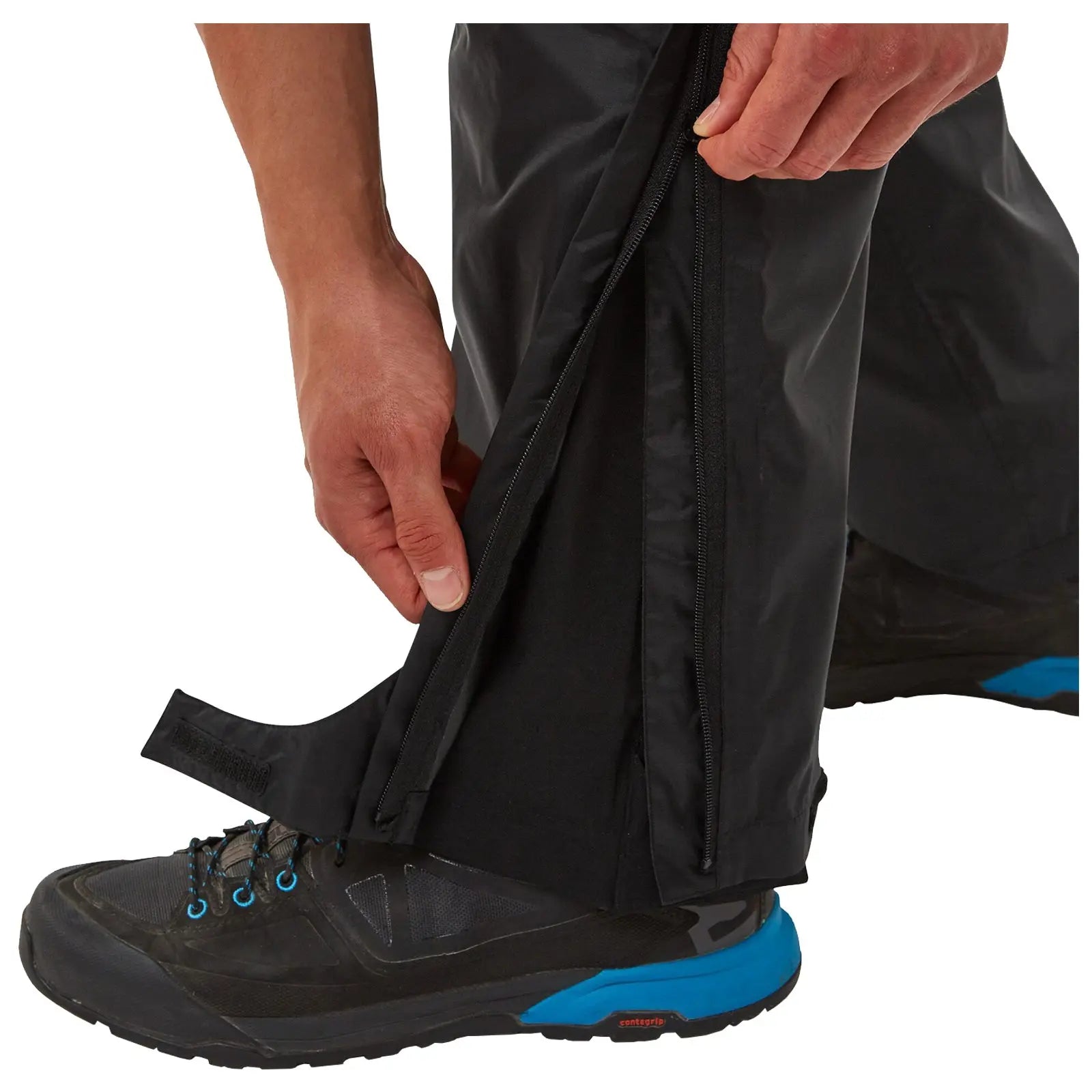 A person is pulling a zipper on the side of black waterproof pants while standing in a black and blue hiking shoe on a flat surface, highlighting an outdoor clothing context.