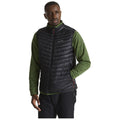 A man is standing with a confident posture wearing a black puffy vest over a green long-sleeve shirt in a neutral background, showcasing a casual outdoor style.