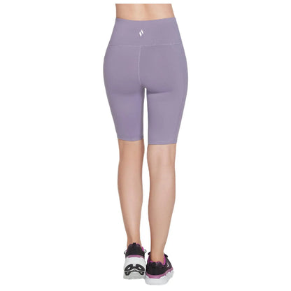 A pair of lavender bike shorts is displayed worn by a person facing away showing a high waistband and fitted design complemented by black and pink athletic shoes in a neutral setting.
