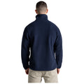 A person is standing with their back facing the viewer wearing a navy blue fleece jacket and beige pants in a plain white background.