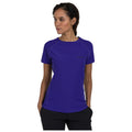 A woman wearing a purple athletic t-shirt stands with her head slightly turned looking to the side in a neutral background, showcasing the shirt's design and fit.