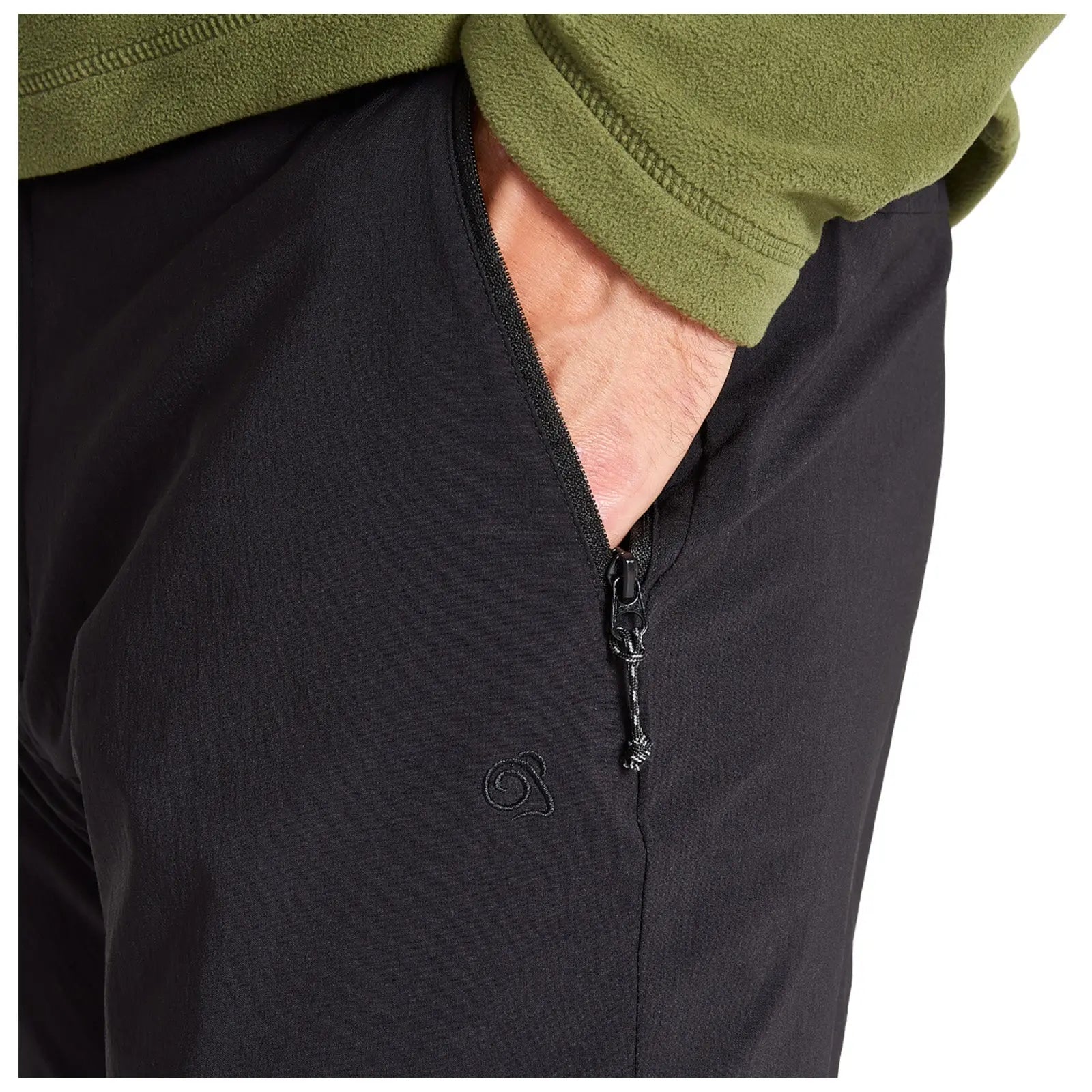 A hand reaches into a zippered pocket of black pants while wearing a green fleece top, showcasing a casual and functional outfit. The pocket features a small logo.
