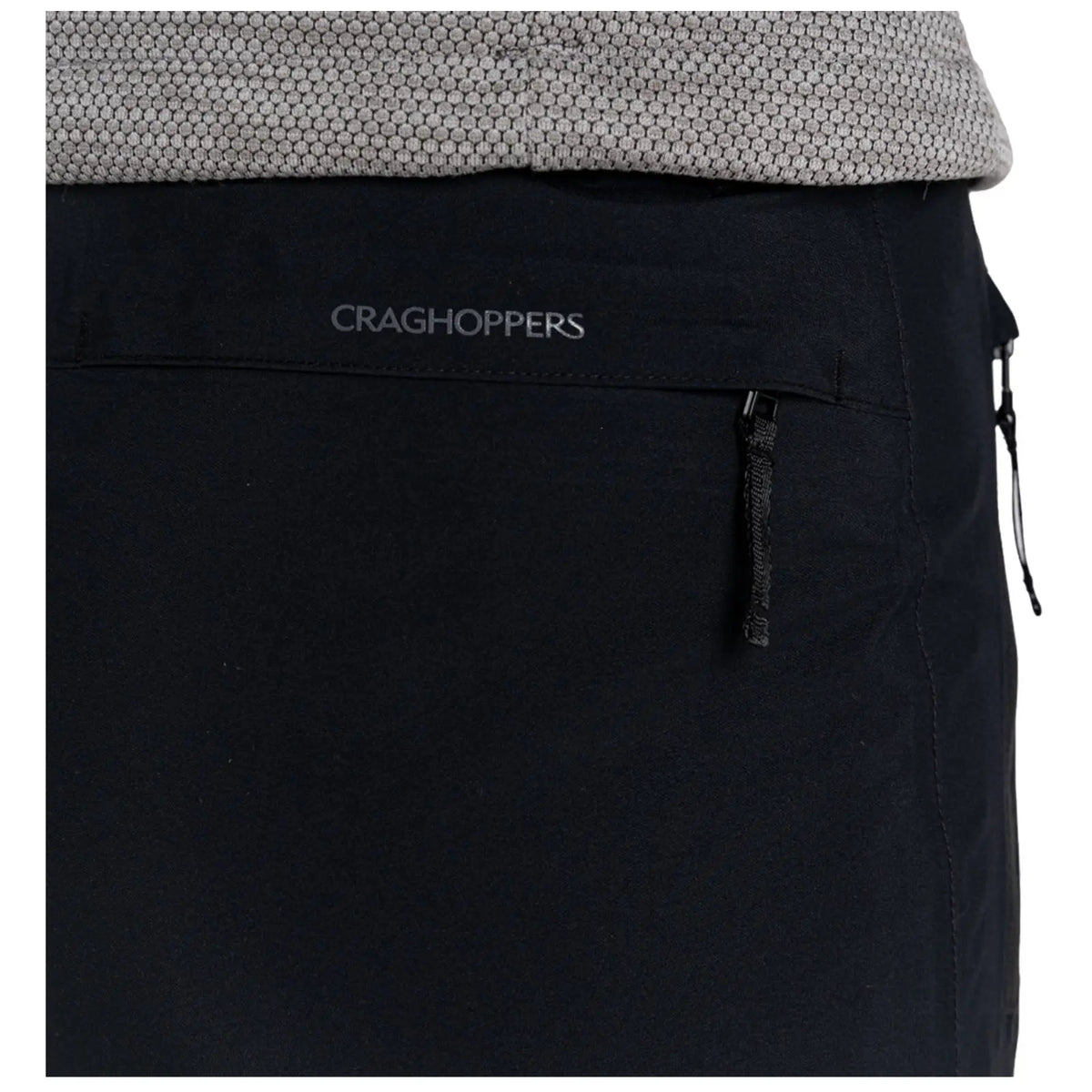 A pair of black pants is displayed with a visible zipper pocket featuring the brand name Craghoppers above it, surrounded by a textured gray fabric at the waist.